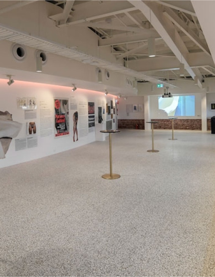 The Ian Pamela Wall Gallery, with a polished concrete floor, small tall bar tables, seating alongside the window and an exhibition on display on the back wall of the room